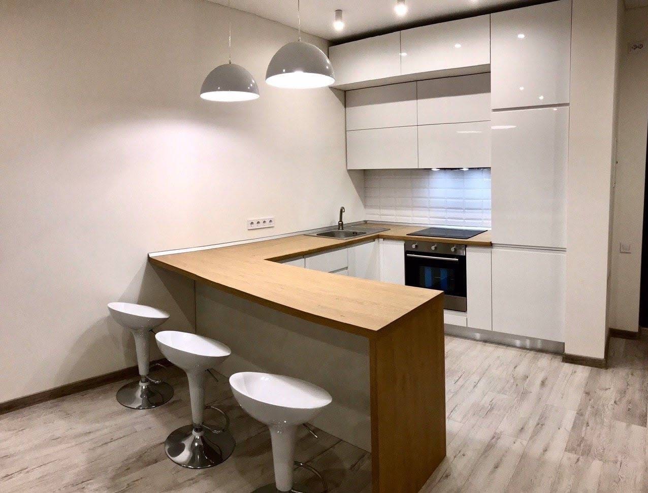 Nowoczesna kuchnia z białymi, lakierowanymi szafkami, drewnianym blatem i trzema białymi hokerami przy wyspie. Minimalistyczne oświetlenie w postaci dwóch okrągłych lamp wiszących.