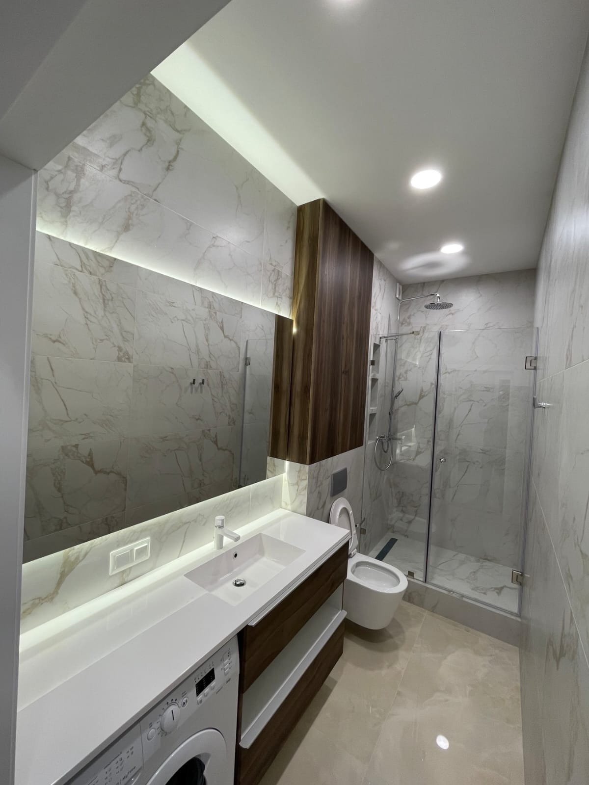 Nowoczesna łazienka z marmurowymi płytkami, podświetlanym lustrem, zabudową z ciemnego drewna, białą umywalką nablatową, toaletą podwieszaną i kabiną prysznicową ze szklanymi drzwiami. Pralka...