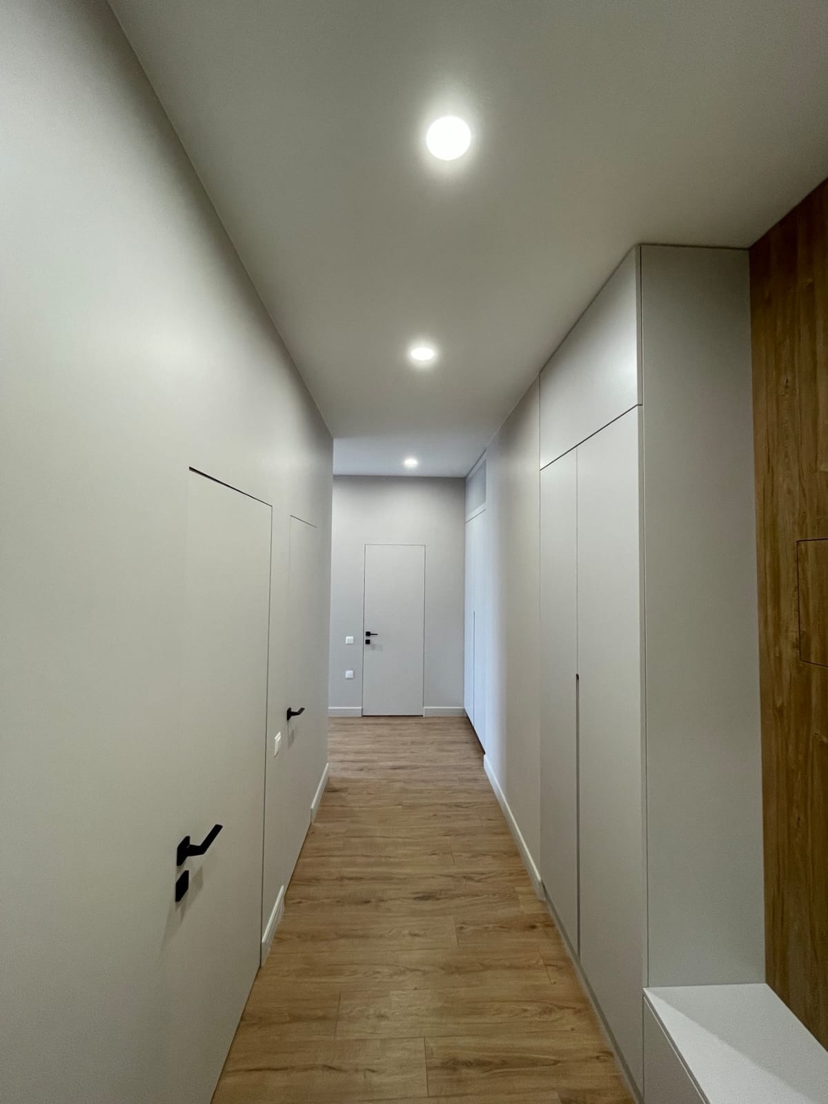 Nowoczesny korytarz z drewnianą podłogą, szarymi ścianami z ukrytymi drzwiami z czarnymi klamkami i zabudową szaf w kolorze ścian, oświetlony punktowymi lampami sufitowymi.