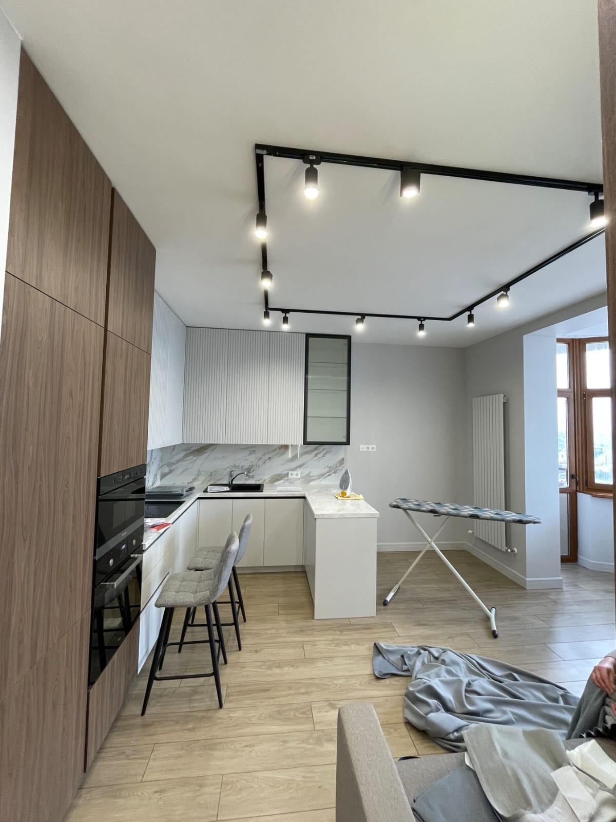 Nowoczesna kuchnia z drewnianymi frontami szafek, białymi szafkami wiszącymi z marmurowym blatem, czarnymi reflektorami na suficie, dwoma szarymi krzesłami barowymi i deską do prasowania