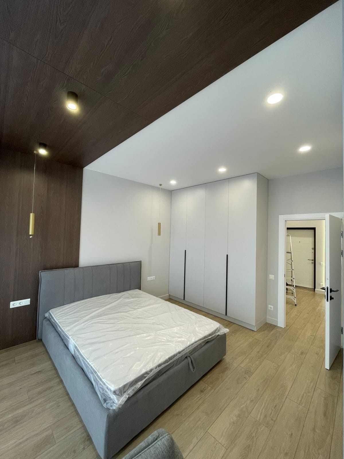 Nowoczesna sypialnia z szarą tapicerowaną ramą łóżka, materacem w folii ochronnej, drewnianą okładziną sufitu i ściany, oraz białą szafą w zabudowie z czarnymi uchwytami, widok na korytarz...