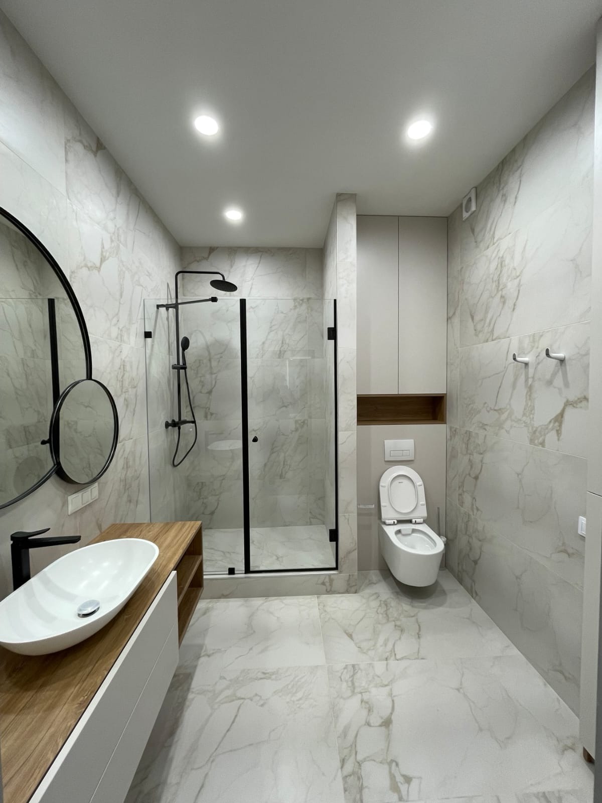 Nowoczesna łazienka z marmurowymi płytkami, kabiną prysznicową typu walk-in z czarnymi ramami, owalną umywalką nablatową na drewnianym blacie i podwieszaną toaletą.