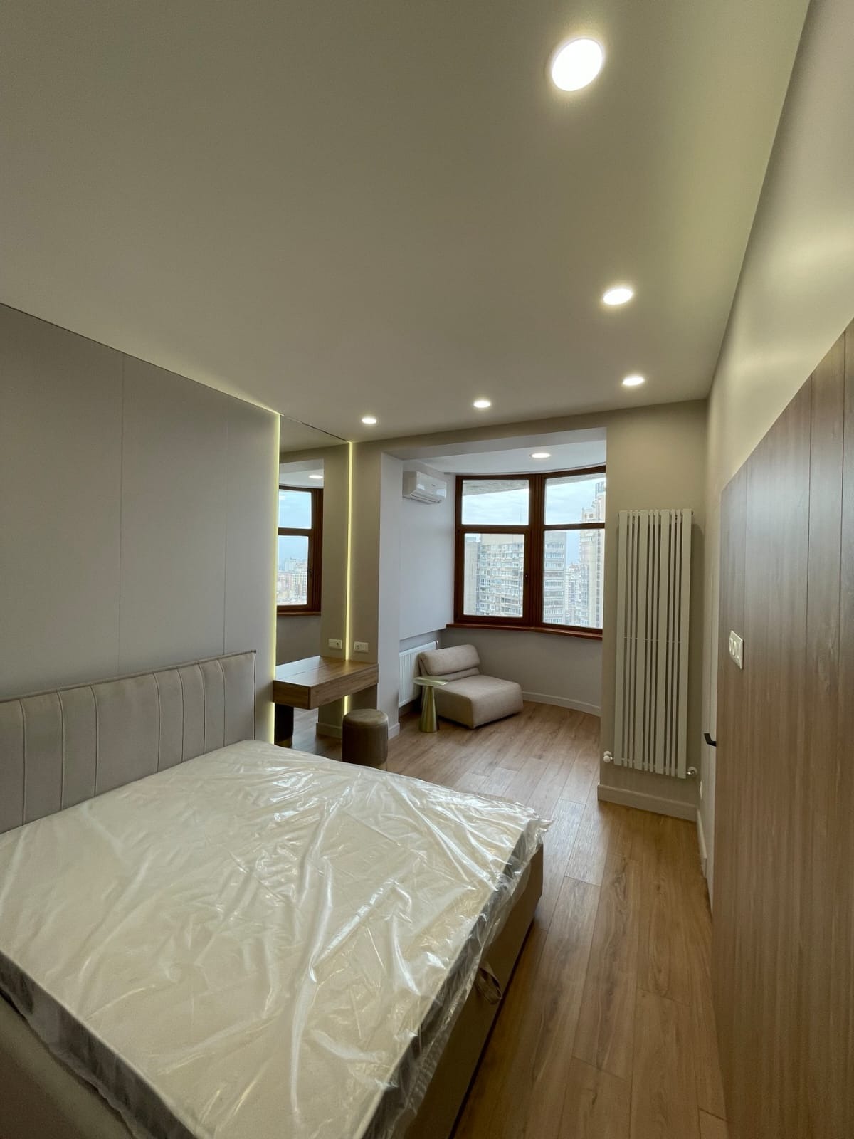 Jasna sypialnia po remoncie z łóżkiem zabezpieczonym folią, drewnianą podłogą, grzejnikiem żeberkowym, toaletką z lustrem i widokiem na miasto z okna.