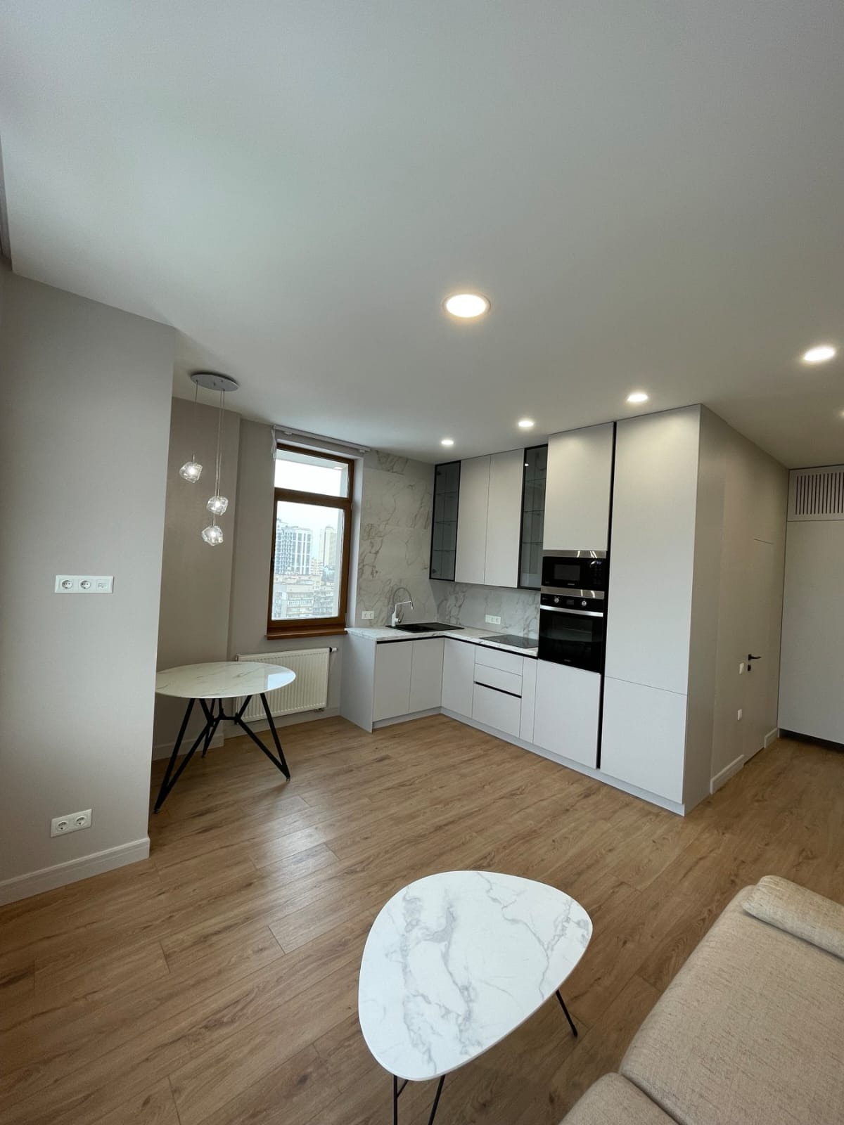 Jasne, nowoczesne wnętrze mieszkania z drewnianą podłogą, białymi meblami kuchennymi z czarnymi akcentami, marmurowym blatem i okrągłym stolikiem z marmurowym blatem na czarnych nogach. Widoczne...