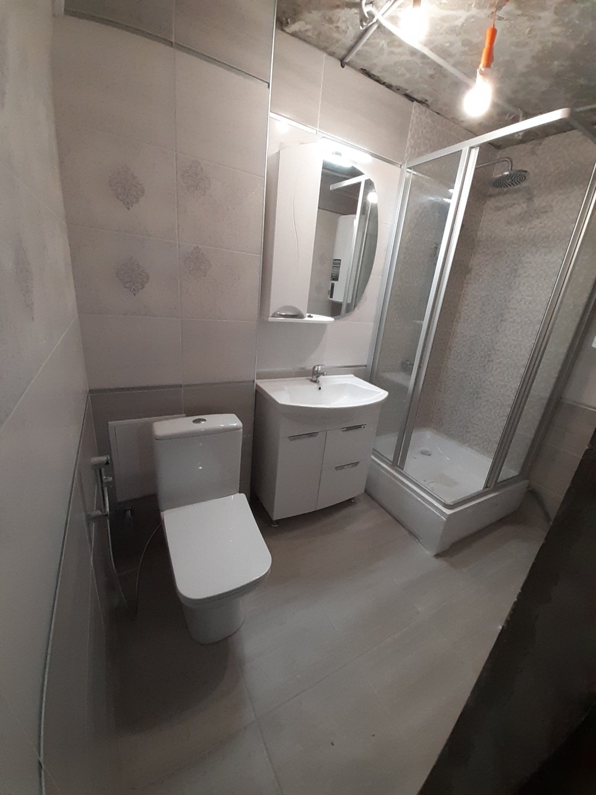 Wykończona łazienka z białą armaturą, kabiną prysznicową z szarymi płytkami i niedokończonym sufitem z widocznymi kablami i żarówką.