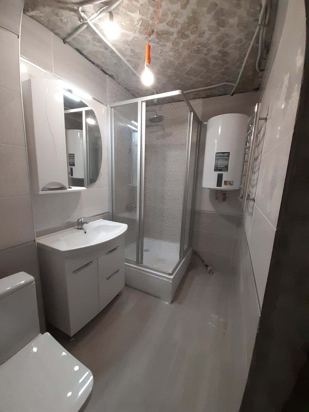 Wykończona łazienka z kabiną prysznicową, umywalką z szafką, grzejnikiem drabinkowym i bojlerem, surowy betonowy sufit z prowizorycznym oświetleniem.