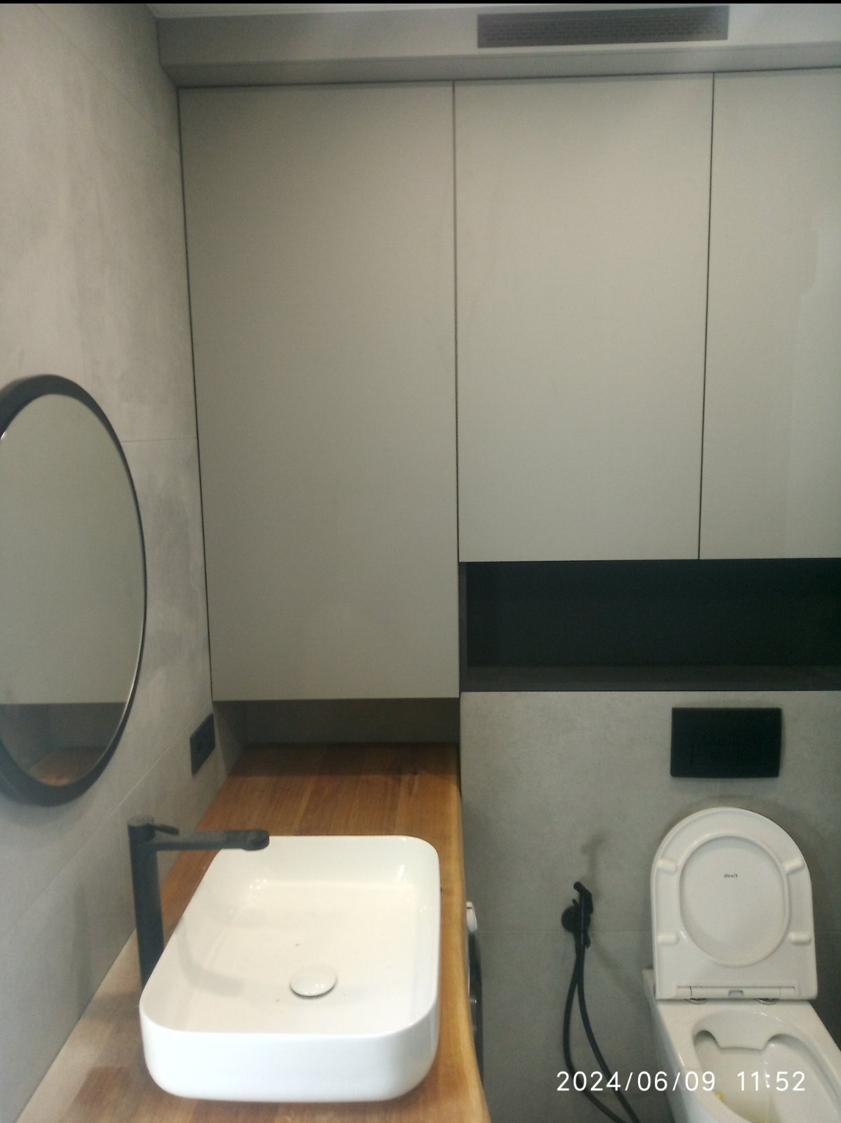 Nowoczesna łazienka z białym umywalką nablatową na drewnianym blacie, okrągłe lustro w czarnej ramie, wisząca toaleta i szare płytki na ścianach.