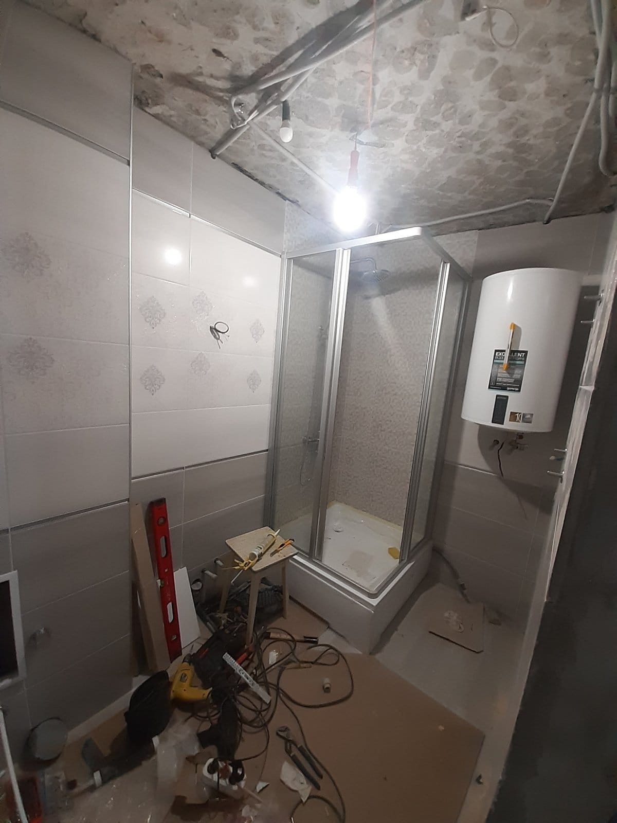 Łazienka w trakcie remontu: kabina prysznicowa z brodzikiem, bojler elektryczny na ścianie z szarymi i białymi płytkami, surowy betonowy sufit z wystającymi kablami, narzędzia i materiały budowlane...