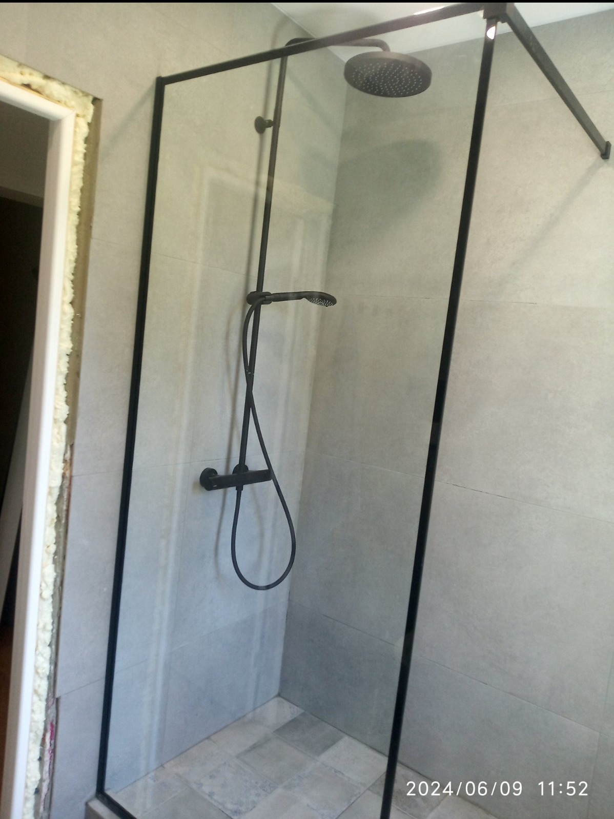 Nowoczesna kabina prysznicowa z czarnymi profilami i deszczownicą, szare płytki na ścianach, widoczna pianka montażowa wokół ościeżnicy drzwi.