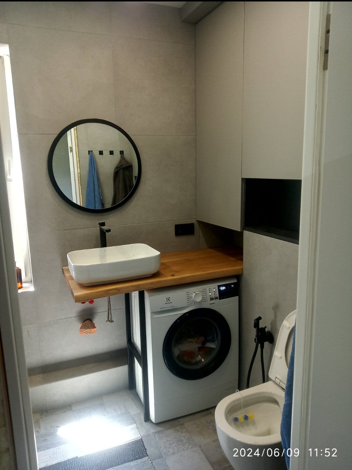 Nowoczesna łazienka z umywalką nablatową na drewnianym blacie wspartym na metalowej konstrukcji nad pralką Electrolux, okrągłe lustro w czarnej ramie, szare płytki na ścianach.