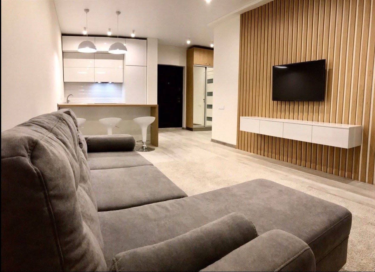 Nowoczesny salon z szarą sofą, drewnianą ścianą z telewizorem i aneksem kuchennym z białymi szafkami oraz barkiem z hokerami.