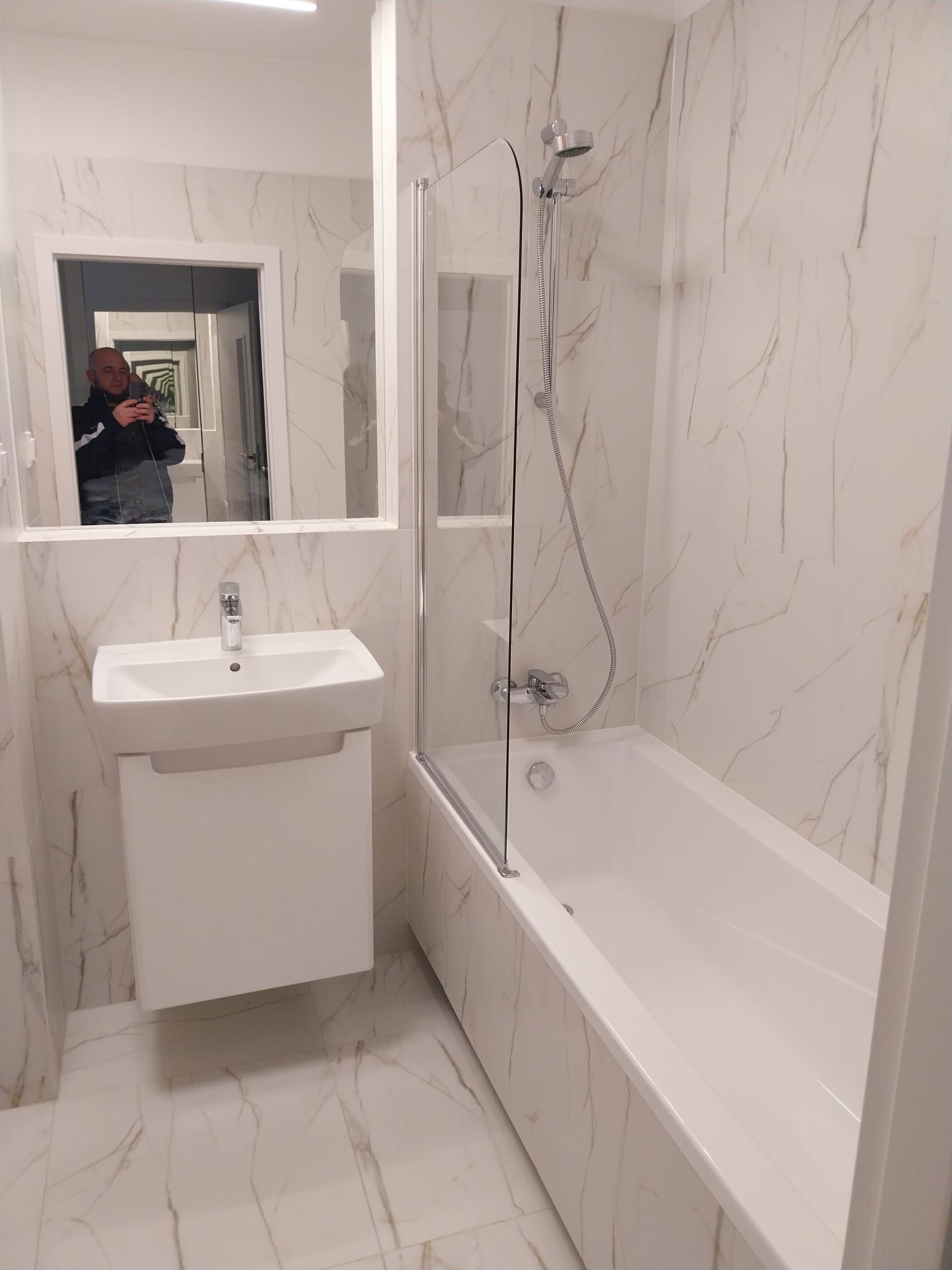 Wykończona łazienka z białymi płytkami imitującymi marmur, białą umywalką z szafką i wanną z parawanem, widoczne odbicie fotografa w lustrze.