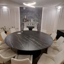 Okrągły stół z blatem imitującym czarny marmur, otoczony kremowymi krzesłami na tle ściany z dekoracyjnym kominkiem i nowoczesną lampą sufitową.