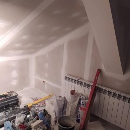 Osoba prywatna - Poddasze w trakcie wykańczania: ściany i skosy pokryte płytami gipsowo-kartonowymi, widoczne narzędzia, poziomica, worki z gipsem i wiadro, grzejnik na ścianie.