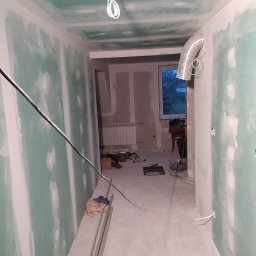 Osoba prywatna - Korytarz w trakcie remontu: ściany i sufit pokryte płytami gipsowo-kartonowymi, widoczne przewody elektryczne, wentylacja w tle, narzędzia i materiały budowlane na podłodze.