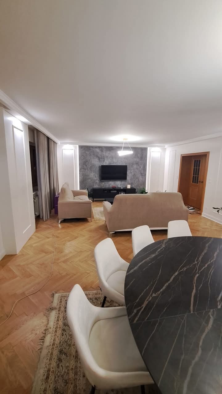 Jasne wnętrze salonu z parkietem w jodełkę, beżowymi sofami, ścianą w odcieniach szarości za telewizorem i okrągłym stołem z blatem imitującym marmur w pierwszym planie.