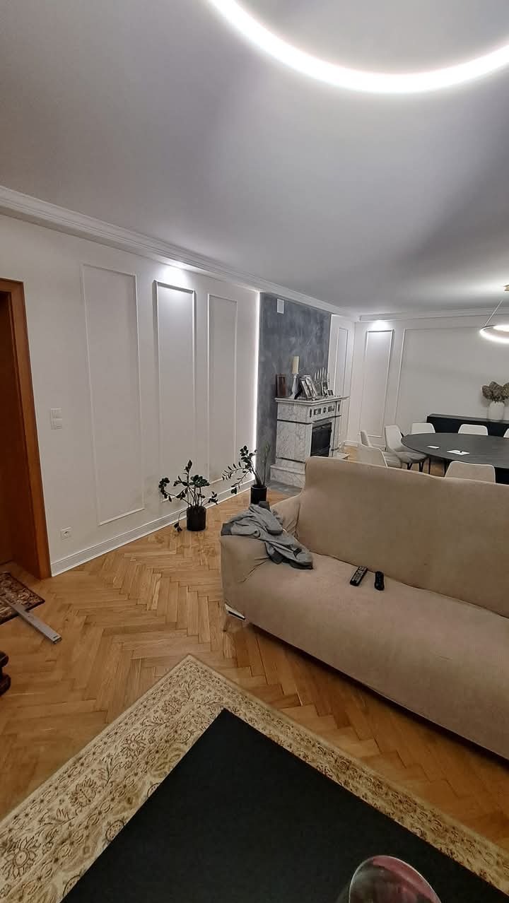 Eleganckie wnętrze salonu z dekoracyjnymi panelami ściennymi, parkietem w jodełkę i podświetleniem LED na suficie. Widoczny marmurowy kominek, sofa z pilotami i rośliny doniczkowe.