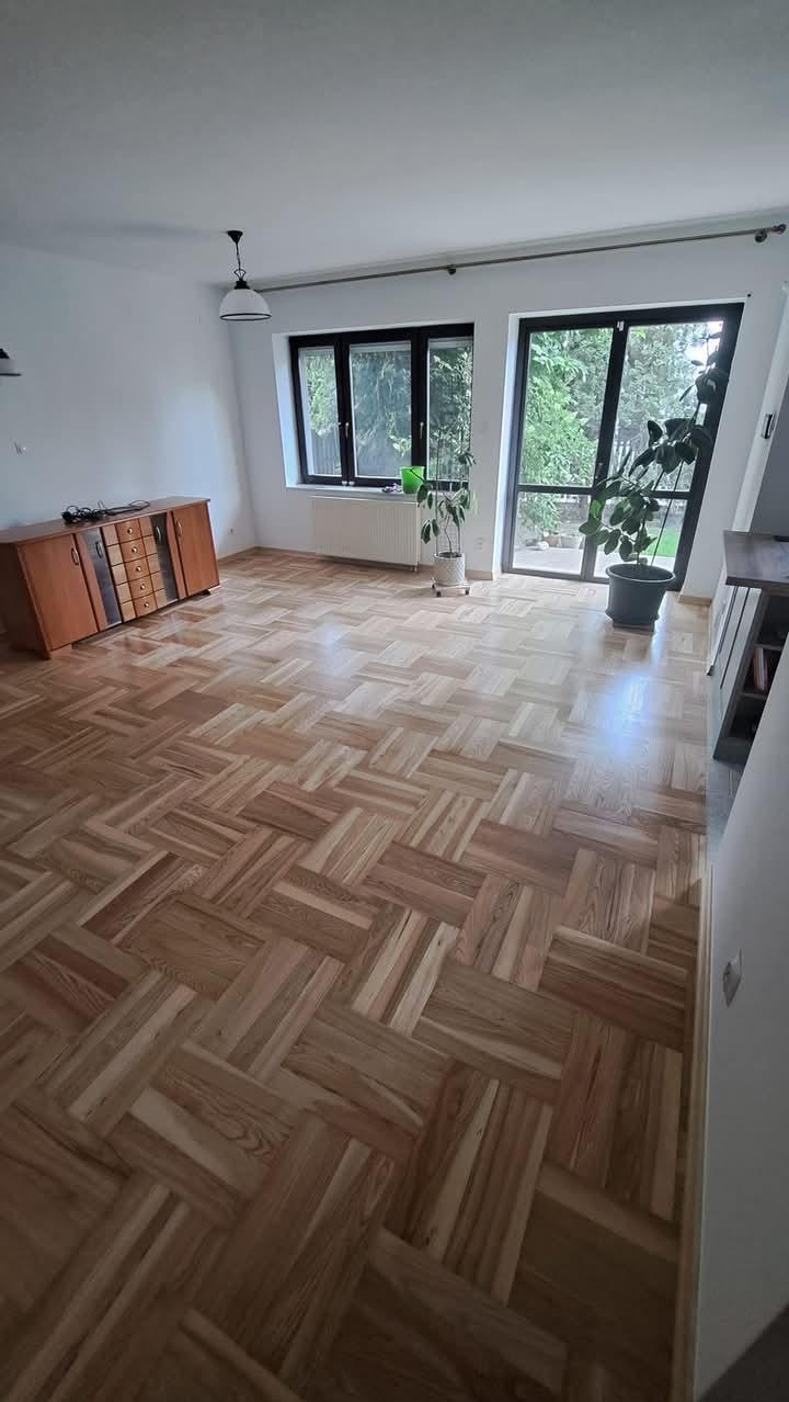 Parkiet ułożony w jodełkę francuską w przestronnym pokoju z dużymi oknami i roślinami doniczkowymi, widoczna komoda z szufladami.