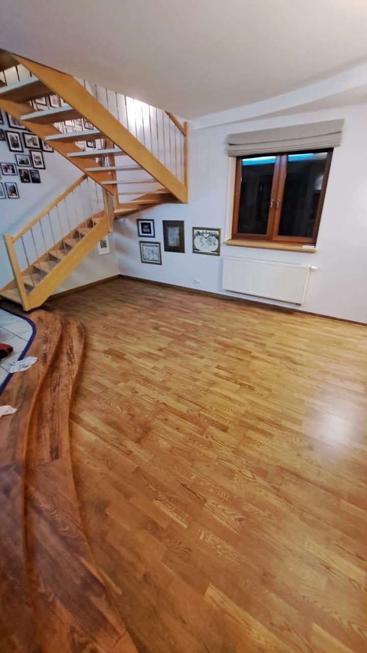 Jasne drewniane schody z białymi balustradami w przestronnym pokoju z parkietem i oknem z roletą rzymską.