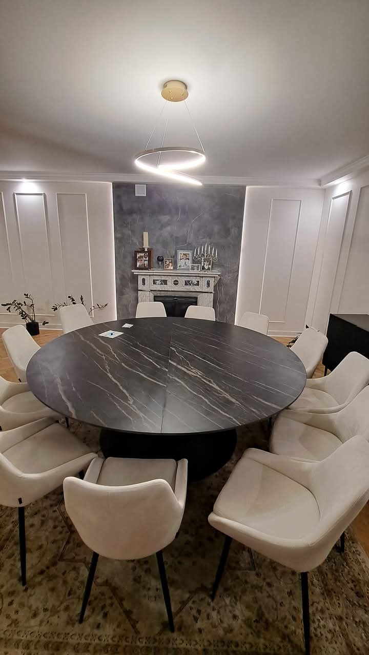 Okrągły stół z blatem imitującym czarny marmur, otoczony kremowymi krzesłami na tle ściany z dekoracyjnym kominkiem i nowoczesną lampą sufitową.