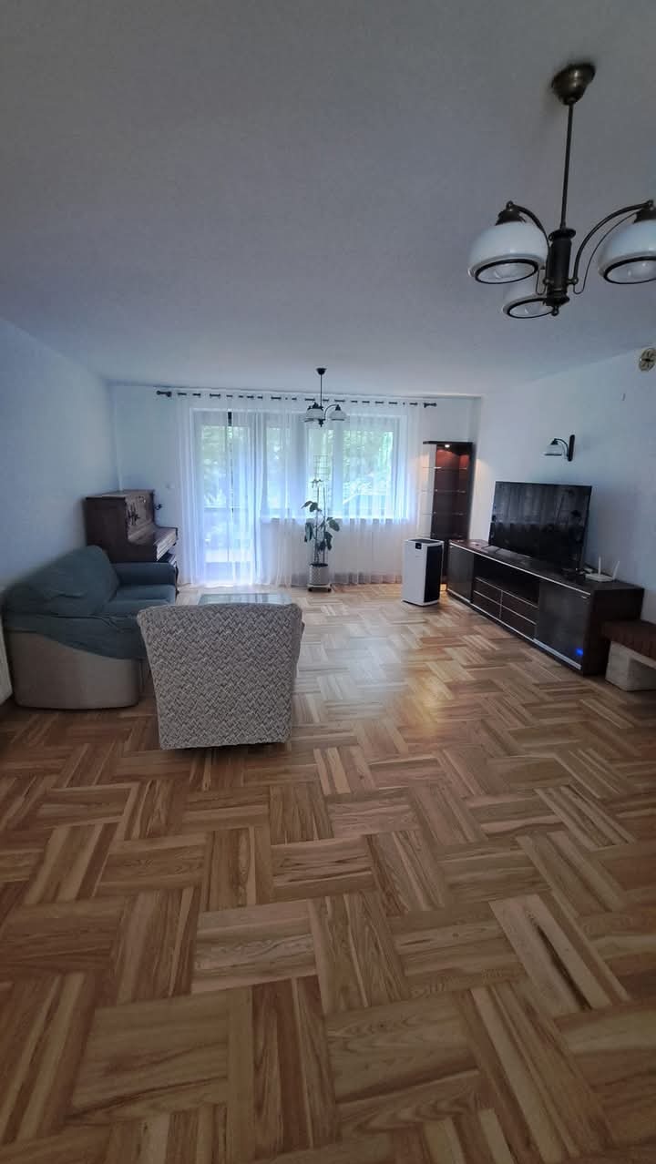 Salon z parkietem ułożonym we wzór jodełki, widok na okno z zasłonami, pianino, kanapę i meble RTV w odcieniach brązu.