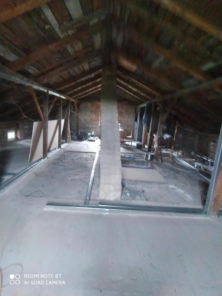 Poddasze w trakcie adaptacji: surowy betonowy podkład, metalowe profile konstrukcyjne ułożone wzdłuż ścian, drewniana więźba dachowa i murowany komin centralnie na planie.