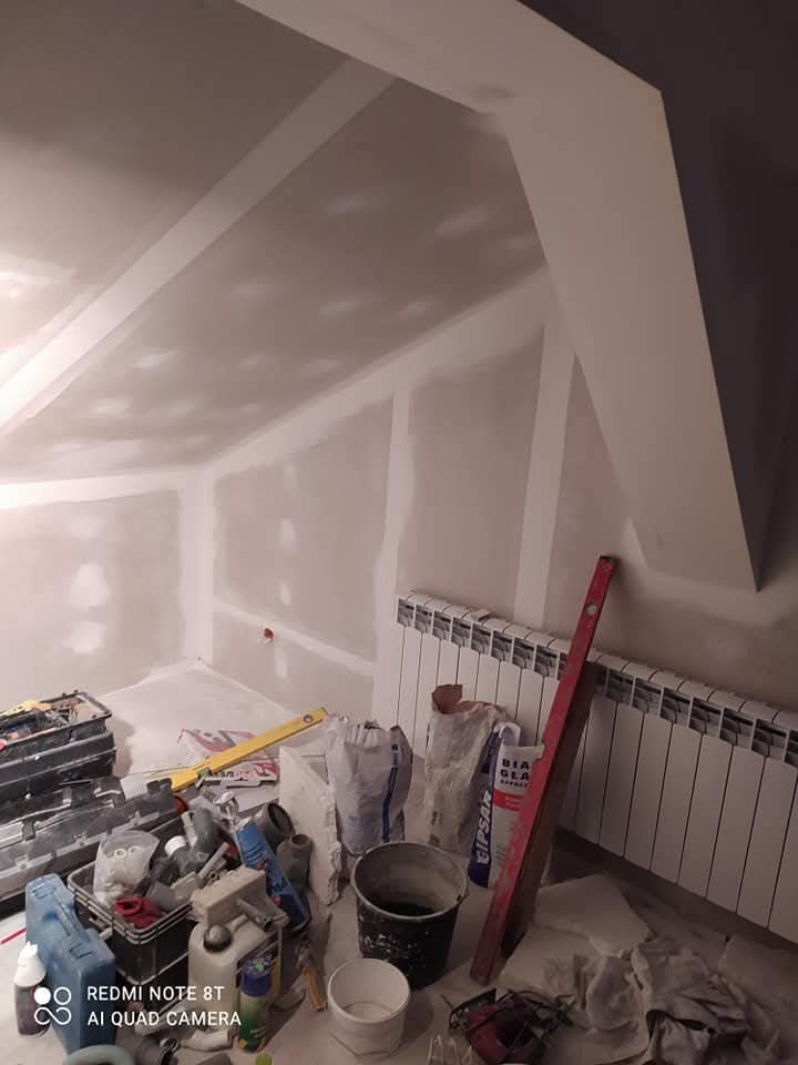 Poddasze w trakcie wykańczania: ściany i skosy pokryte płytami gipsowo-kartonowymi, widoczne narzędzia, poziomica, worki z gipsem i wiadro, grzejnik na ścianie.