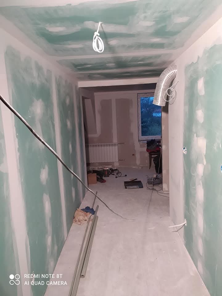 Korytarz w trakcie remontu: ściany i sufit pokryte płytami gipsowo-kartonowymi, widoczne przewody elektryczne, wentylacja w tle, narzędzia i materiały budowlane na podłodze.