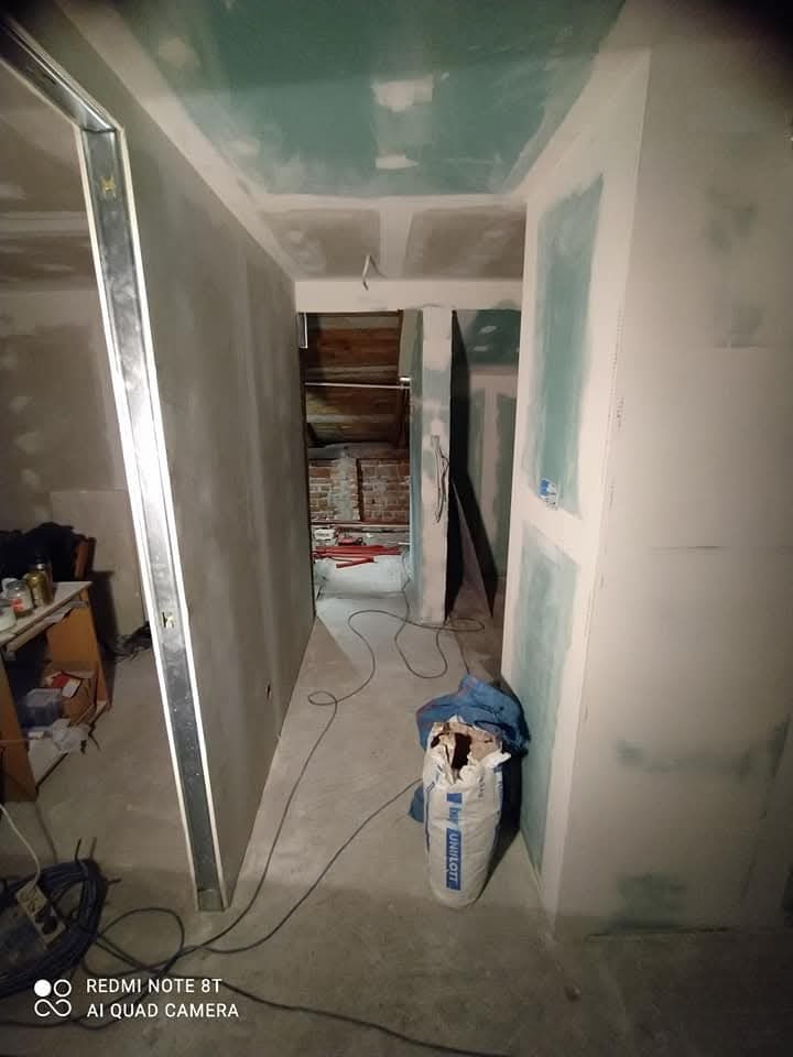 Wnętrze w trakcie remontu: surowy korytarz z widocznymi płytami gipsowo-kartonowymi na ścianach i suficie, metalowe profile, worek z zaprawą Unilott na podłodze oraz częściowo odsłonięta cegła...