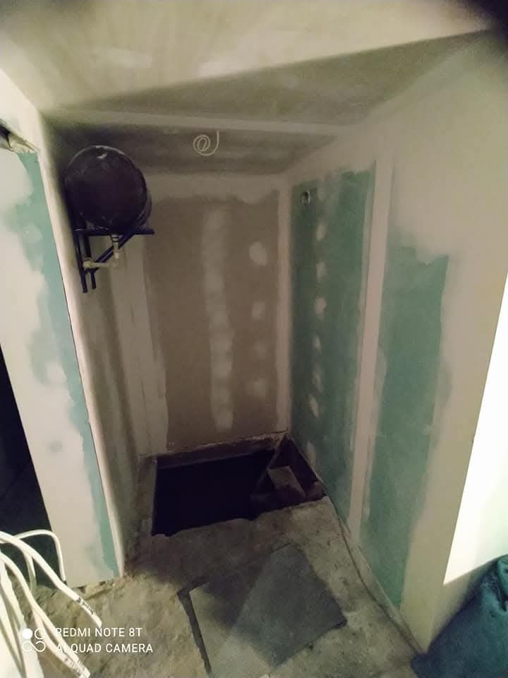 Wnętrze pomieszczenia w trakcie prac wykończeniowych: surowe ściany z widocznymi płytami gipsowo-kartonowymi, niezabezpieczony otwór w podłodze oraz wiszący, okrągły przedmiot na ścianie.