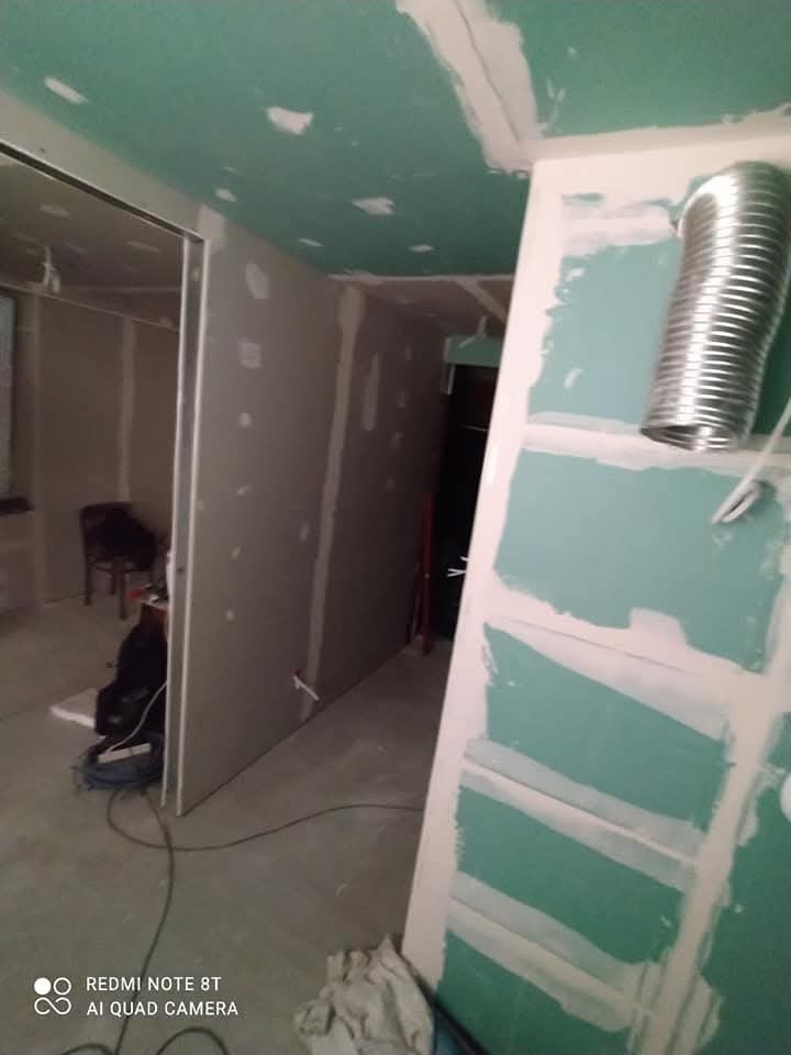 Wnętrze w trakcie remontu: ściany pokryte płytami gipsowo-kartonowymi, częściowo zagruntowane na zielono, widoczna metalowa rura wentylacyjna i przewody na podłodze.