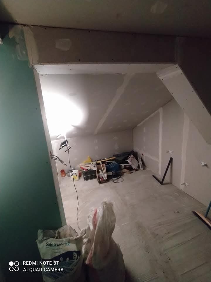 Adaptacja poddasza: surowe ściany pokryte płytami gipsowo-kartonowymi, widoczne łączenia wymagające szpachlowania, podłoga przygotowana pod wylewkę, narzędzia i materiały budowlane w nieładzie.