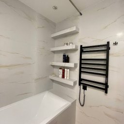 New House - Wykończona łazienka z wanną, grzejnikiem drabinkowym w kolorze czarnym i trzema półkami z kosmetykami na ścianie wyłożonej płytkami imitującymi marmur.