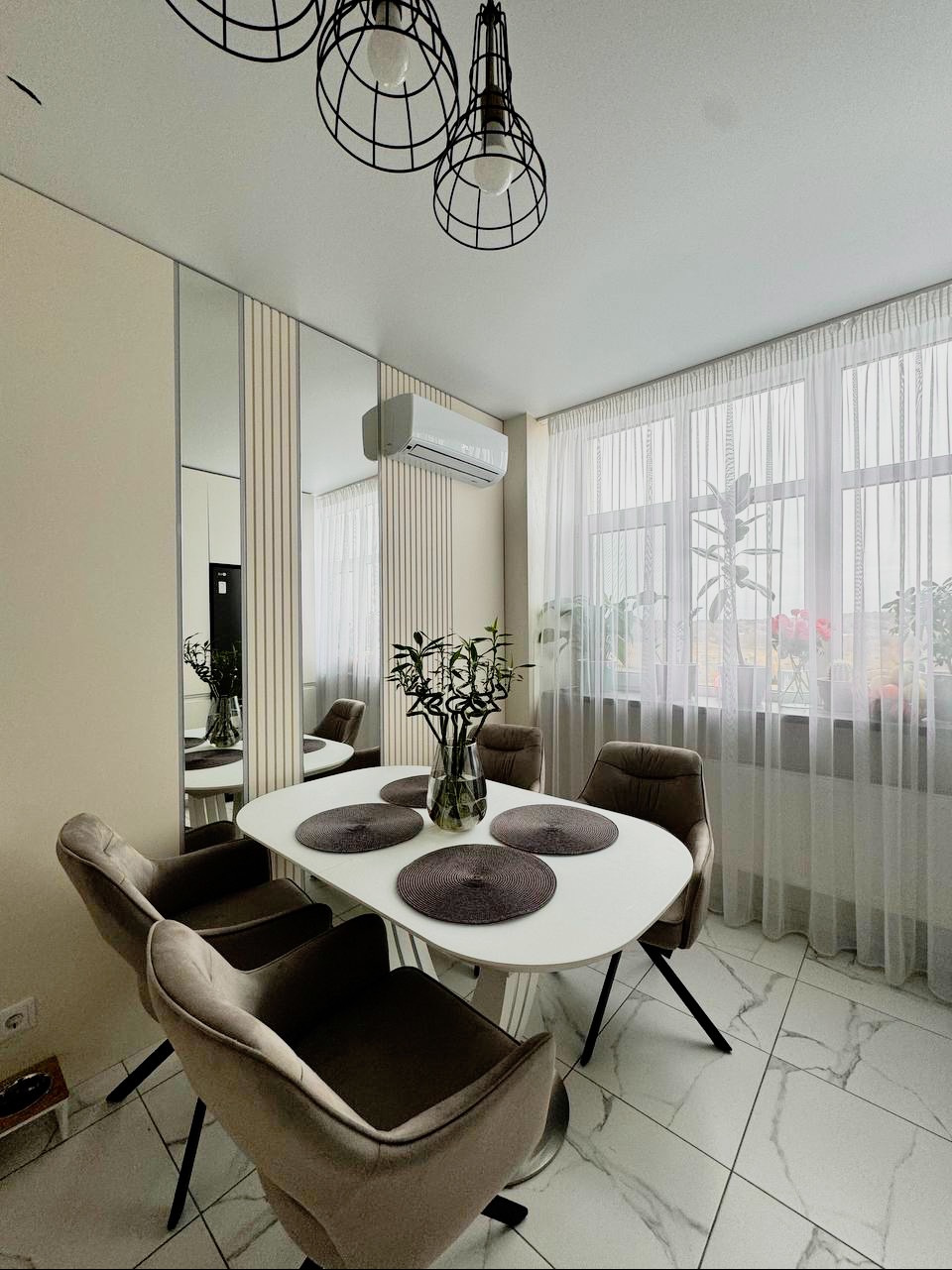 Jasna jadalnia z owalnym białym stołem, czterema szarymi krzesłami, lśniącą podłogą w marmurkowy wzór i lustrami na ścianie, oświetlona trzema wiszącymi lampami w stylu industrialnym.