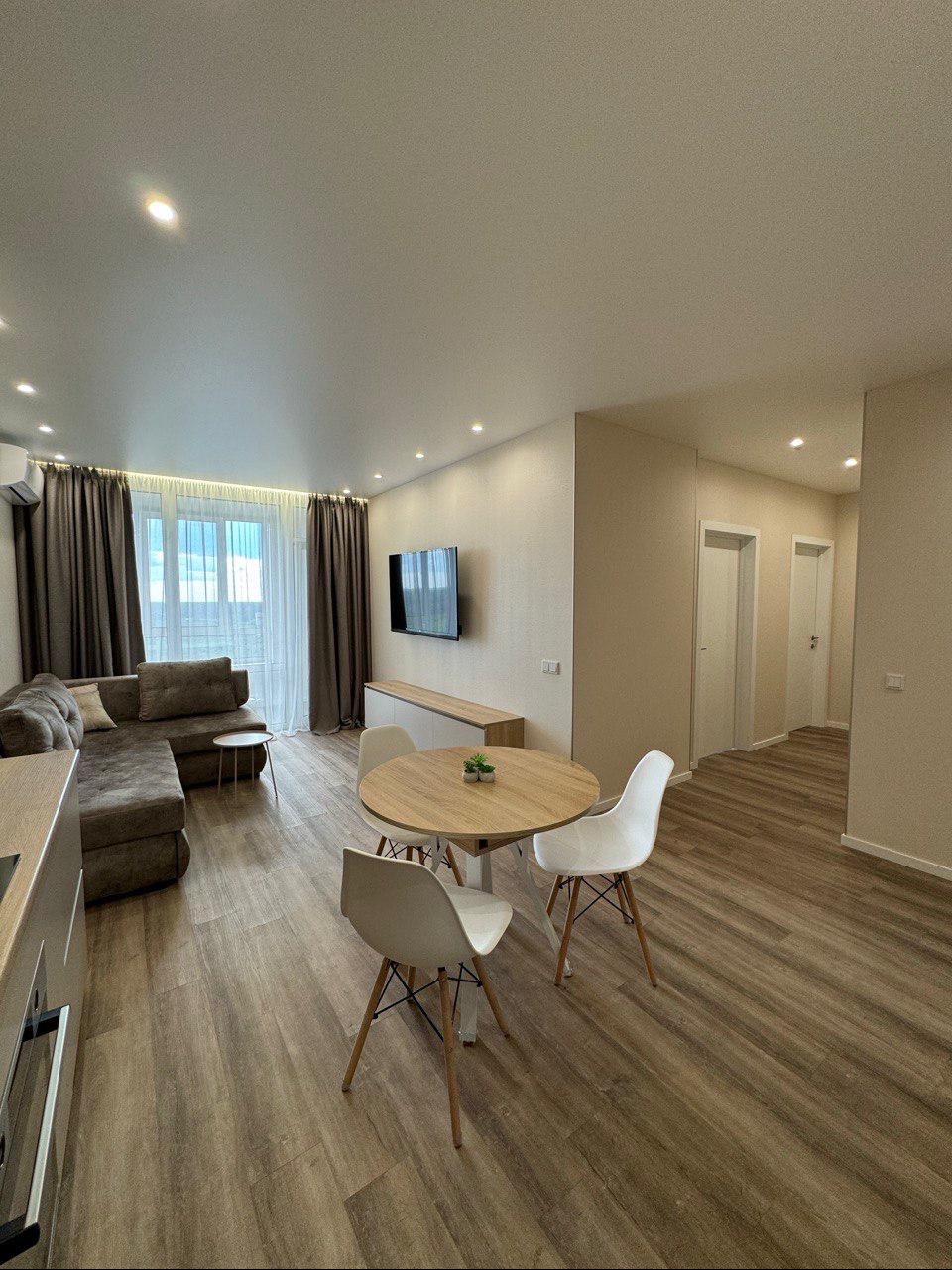 Jasne wnętrze salonu z drewnianą podłogą, brązową sofą, okrągłym stołem z trzema białymi krzesłami i telewizorem na ścianie.