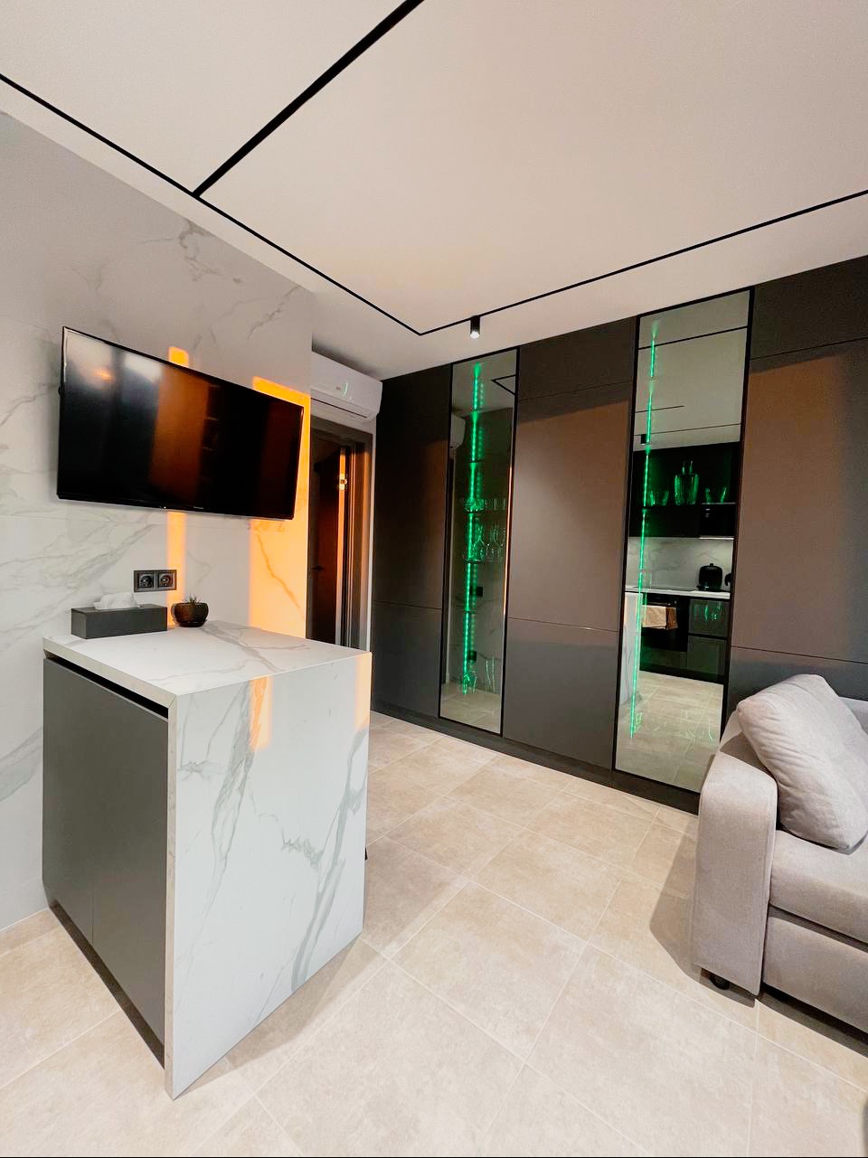 Nowoczesna kuchnia z wyspą o marmurowym blacie, ciemnoszarymi szafkami i lustrzanymi witrynami z zielonym podświetleniem LED, beżowa podłoga z płytek.