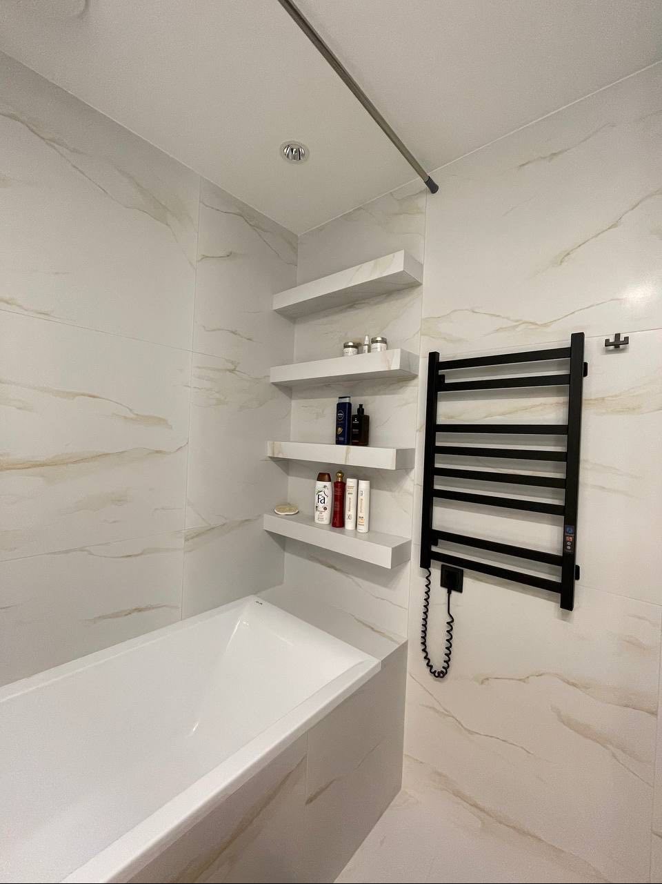 Wykończona łazienka z wanną, grzejnikiem drabinkowym w kolorze czarnym i trzema półkami z kosmetykami na ścianie wyłożonej płytkami imitującymi marmur.