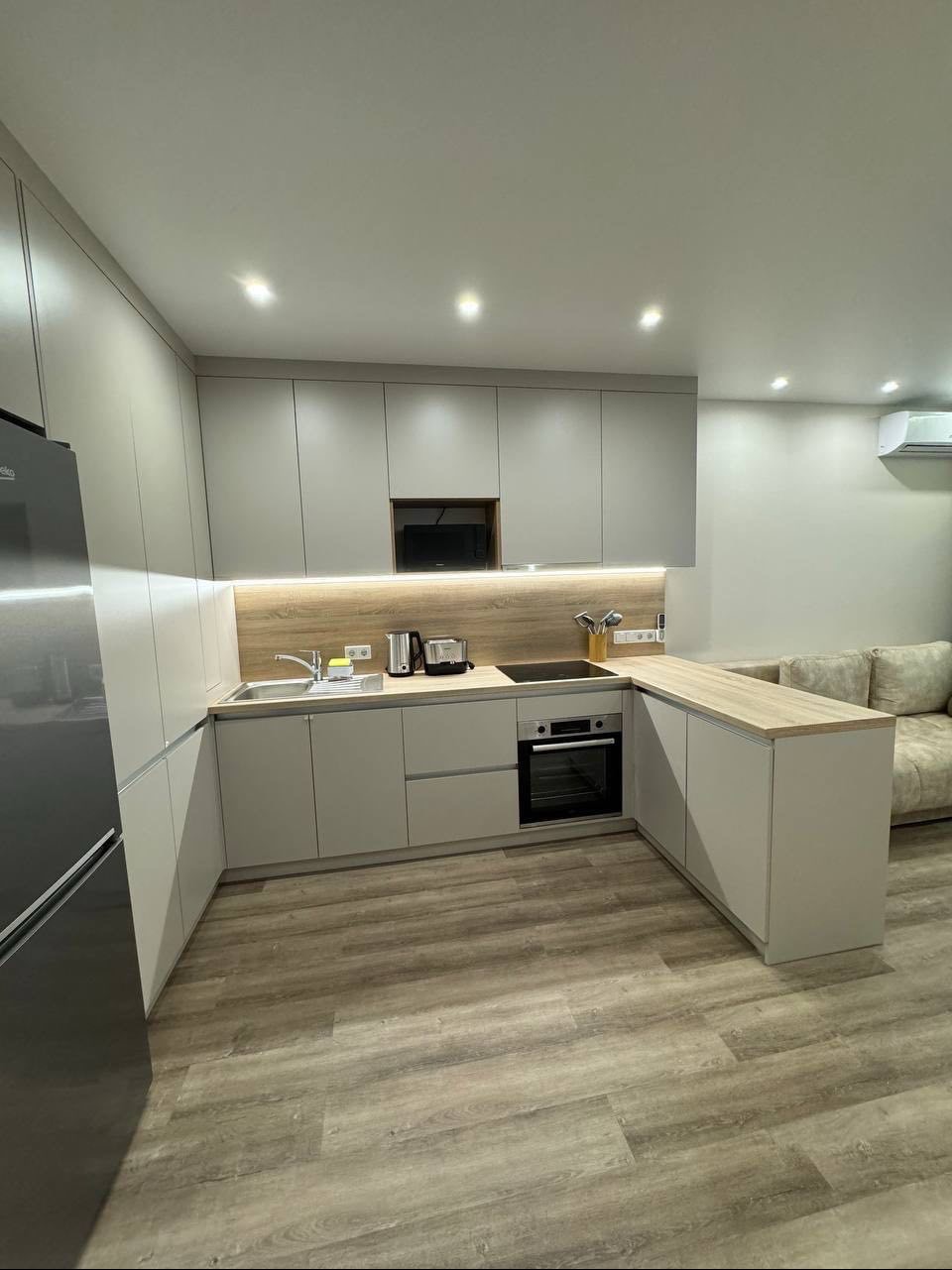 Nowoczesna kuchnia w odcieniach szarości z drewnianymi akcentami, oświetleniem LED pod szafkami i sprzętem AGD, widok na fragment salonu z kanapą w tle.