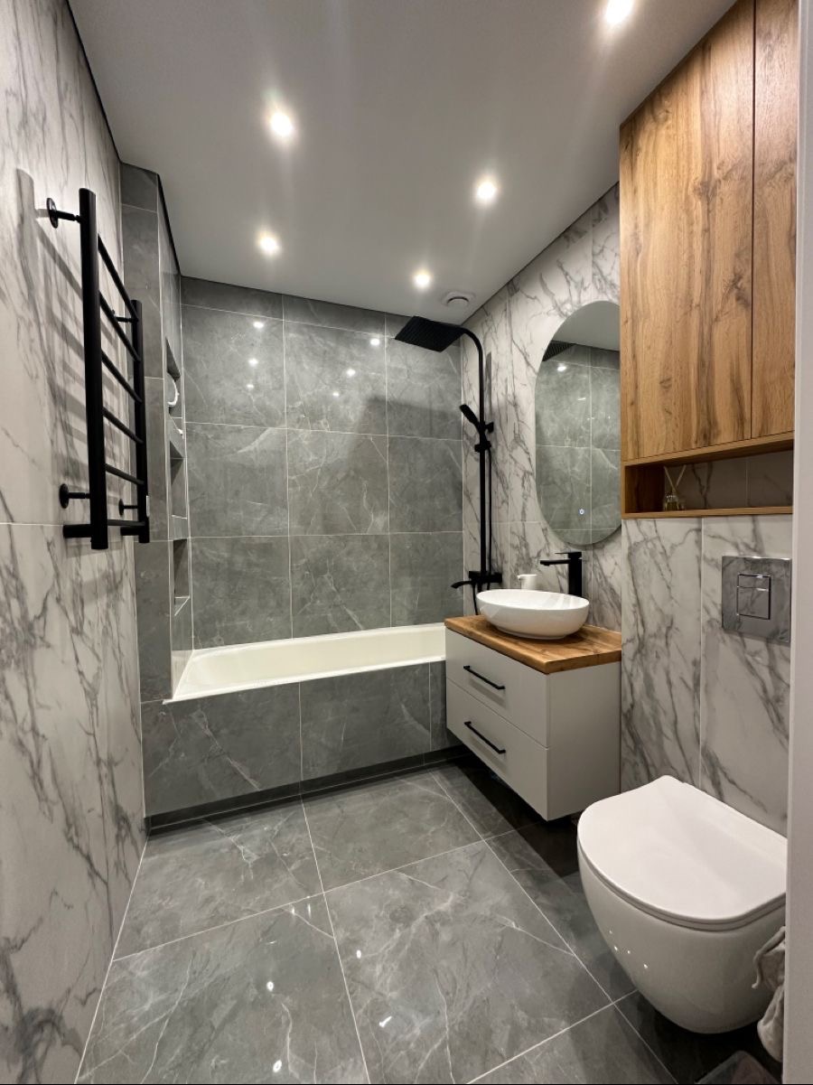 Nowoczesna łazienka w Gliwicach z szarymi płytkami imitującymi marmur, czarną armaturą, owalnym lustrem i drewnianą szafką wiszącą.