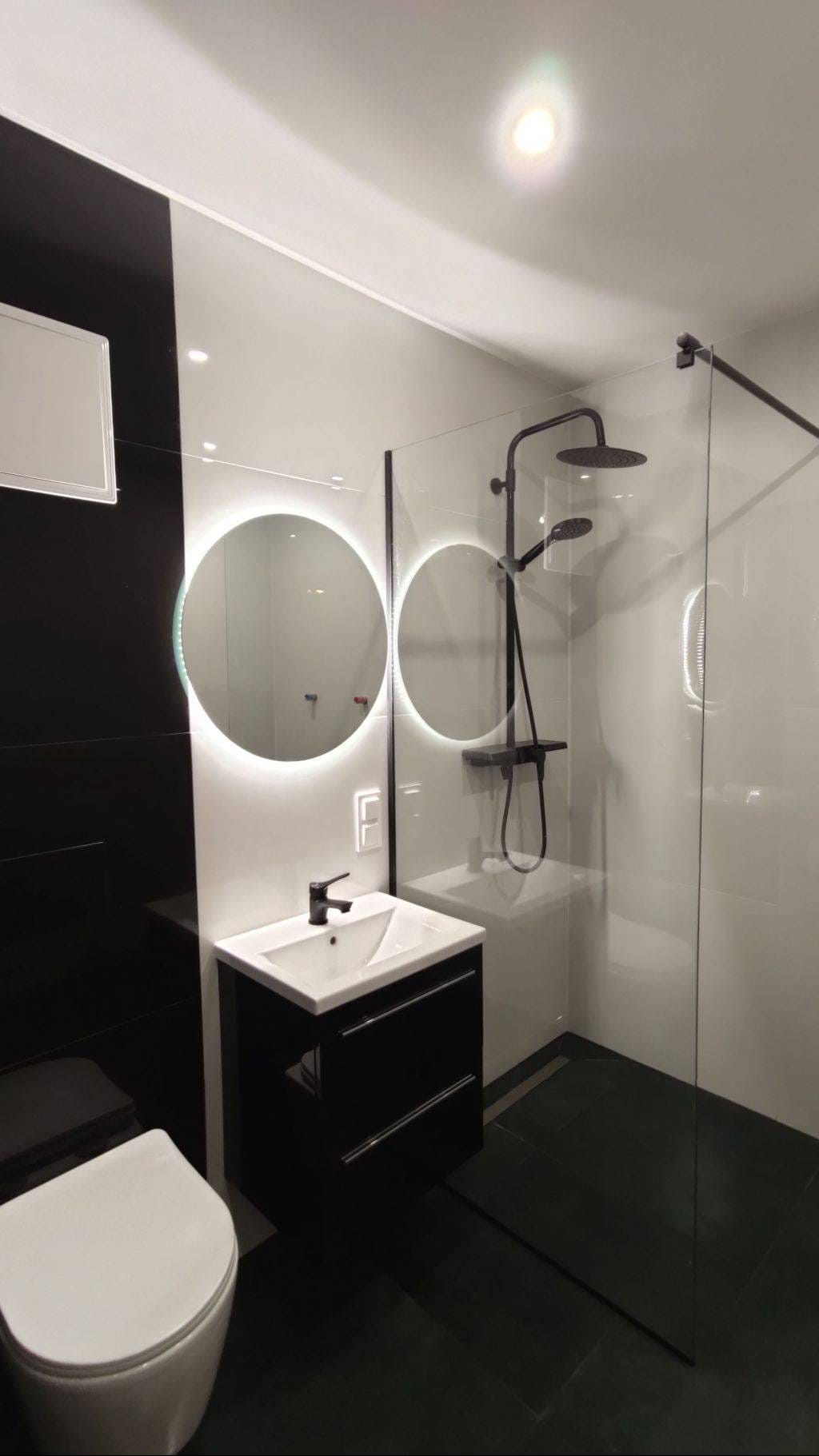 Nowoczesna łazienka w tonacji czerni i bieli z okrągłym lustrem LED, czarną armaturą i kabiną prysznicową typu walk-in z odpływem liniowym w podłodze.