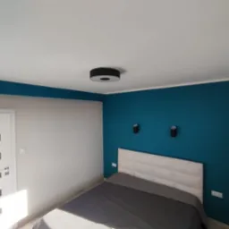 Sypialnia po odświeżeniu: ściana pomalowana na intensywny turkus, biała ściana z drzwiami z poziomymi szarymi pasami, łóżko z szarym nakryciem i białym zagłówkiem, okrągła lampa sufitowa i dwie...