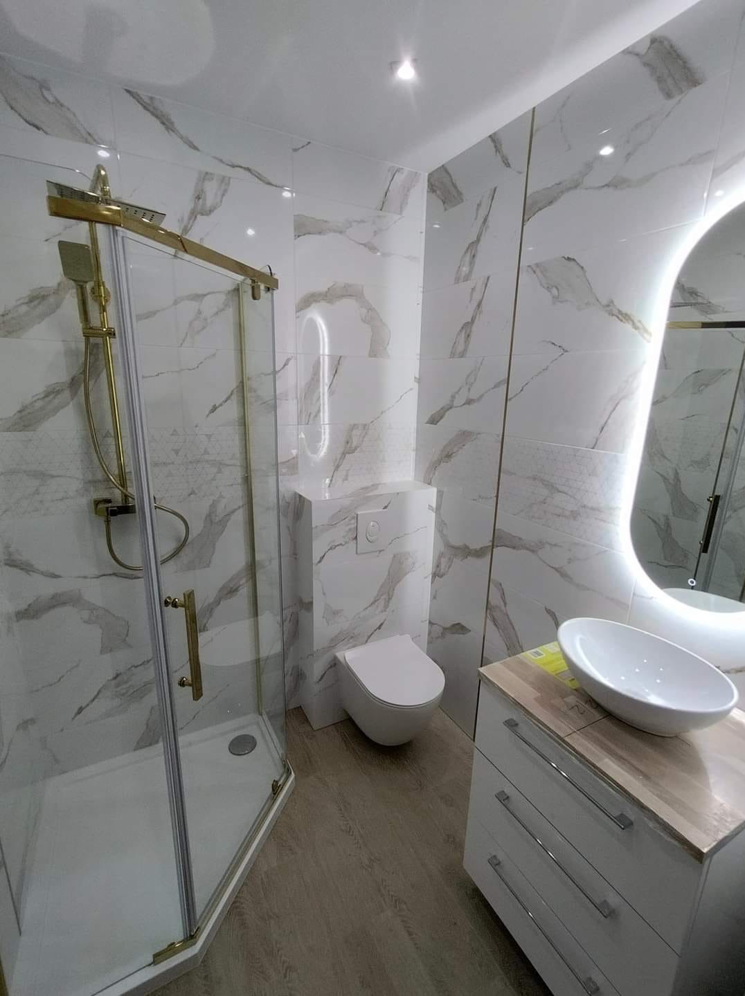 Nowoczesna łazienka z kabiną prysznicową ze złotymi detalami, umywalką nablatową na białej szafce z drewnianym blatem i toaletą w zabudowie, wykończona białymi płytkami imitującymi marmur...