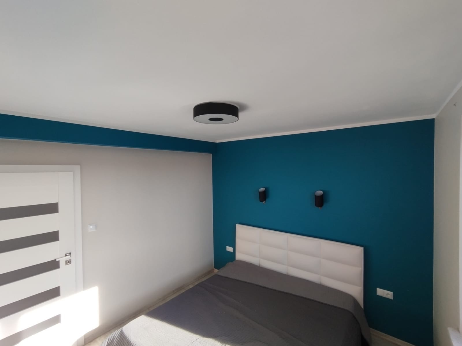 Sypialnia po odświeżeniu: ściana pomalowana na intensywny turkus, biała ściana z drzwiami z poziomymi szarymi pasami, łóżko z szarym nakryciem i białym zagłówkiem, okrągła lampa sufitowa i dwie...