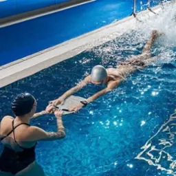 Instruktor pływania asekuruje mężczyznę płynącego z deską w basenie, widoczne pluski wody i błękitna woda.