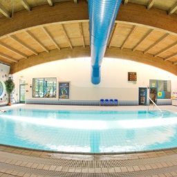 dobrzeplywaj.pl - Panoramiczne ujęcie krytego basenu z jasną wodą, drewnianą konstrukcją dachu i osobami w wodzie; w tle widoczne okna i dekoracje ścienne.