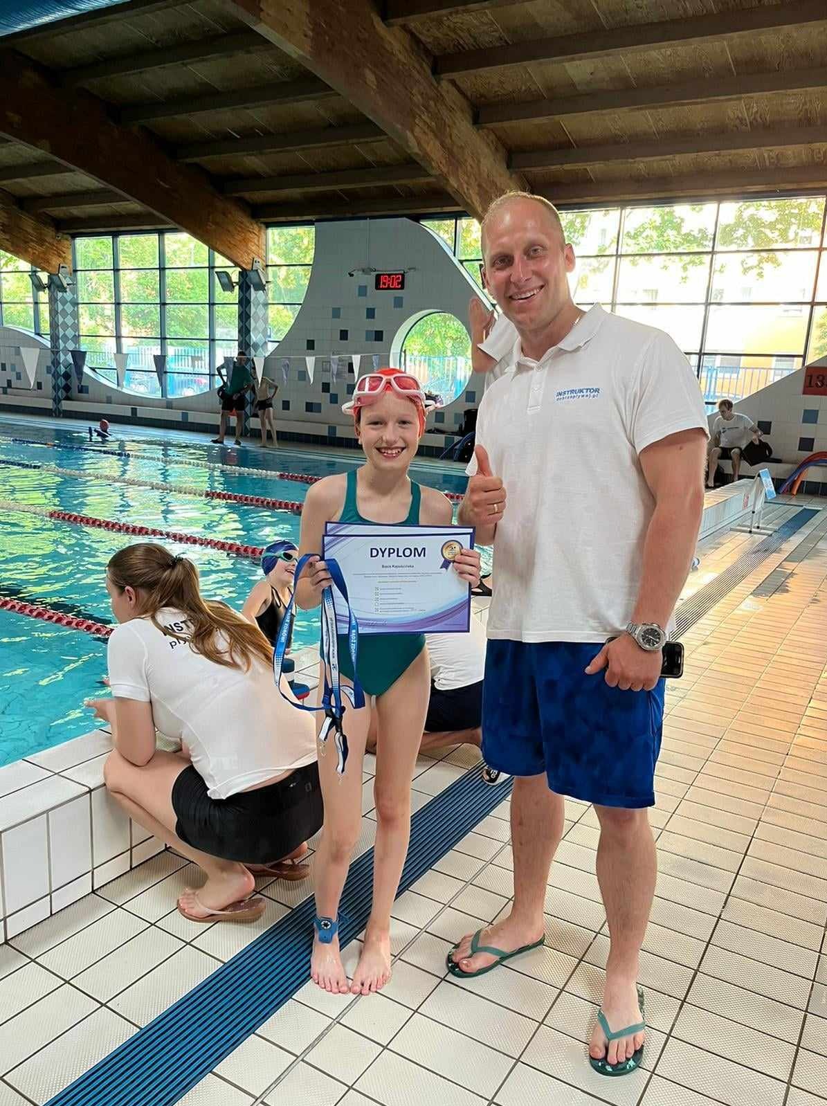 Uśmiechnięta dziewczynka w zielonym stroju kąpielowym i różowych okularach pływackich trzyma dyplom i medale po lekcji pływania, obok stoi instruktor z uniesionym kciukiem, w tle basen.