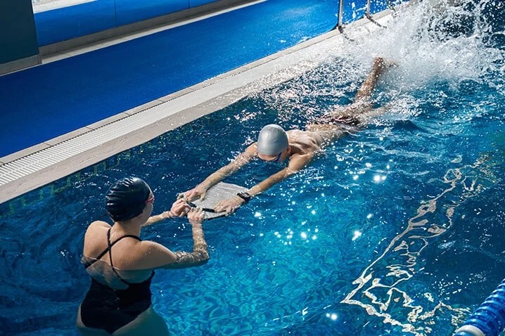 Instruktor pływania asekuruje mężczyznę płynącego z deską w basenie, widoczne pluski wody i błękitna woda.