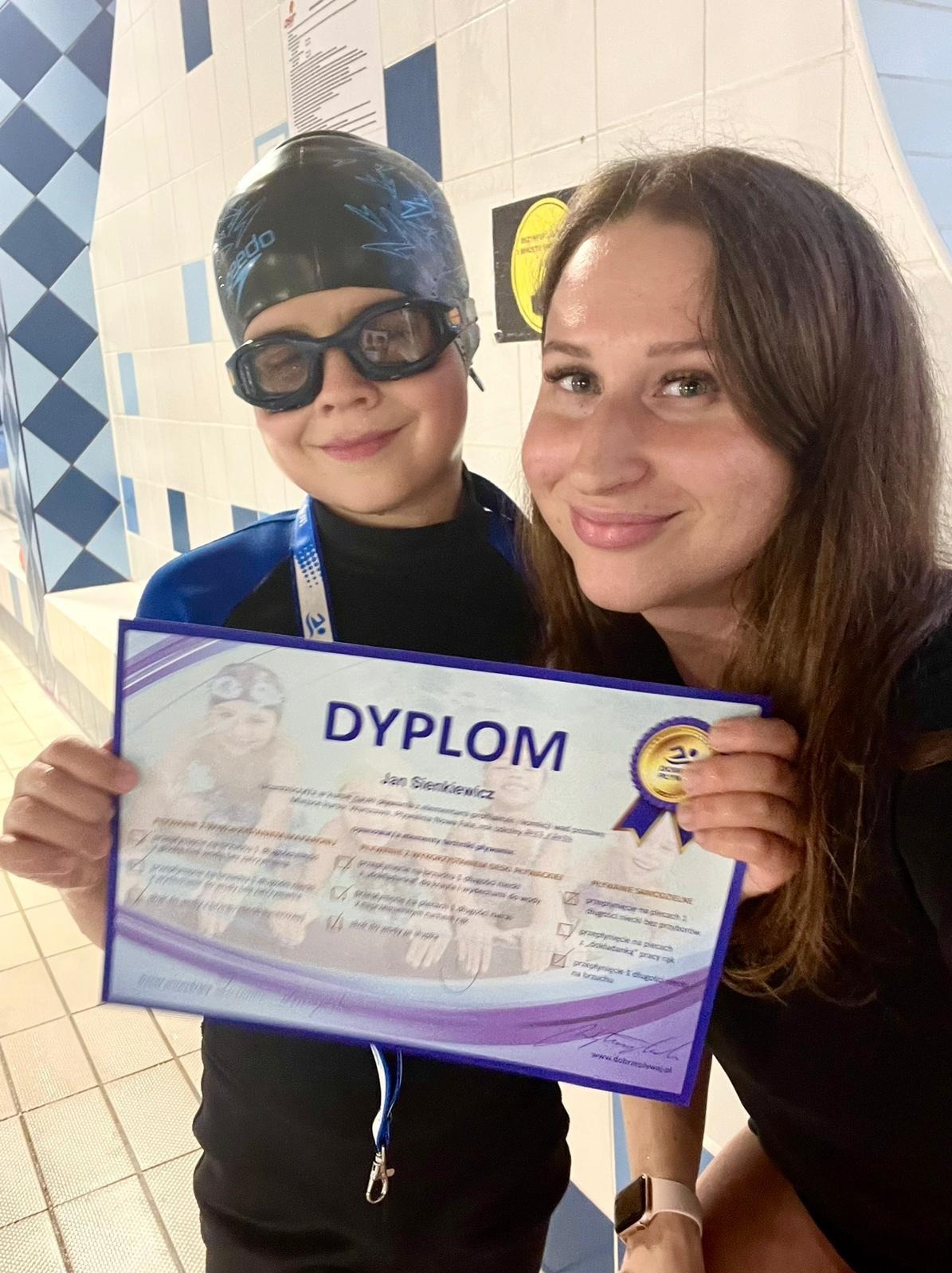 Uśmiechnięty chłopiec w czepku i okularach pływackich, trzymający dyplom ukończenia kursu pływania, obok instruktorka pływania