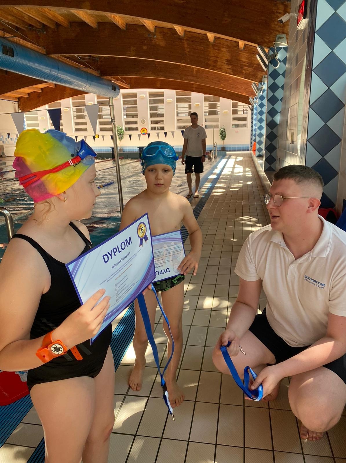 Dwoje dzieci w strojach kąpielowych i czepkach trzyma dyplomy, obok nich instruktor pływania z niebieską smyczą, basen w tle.