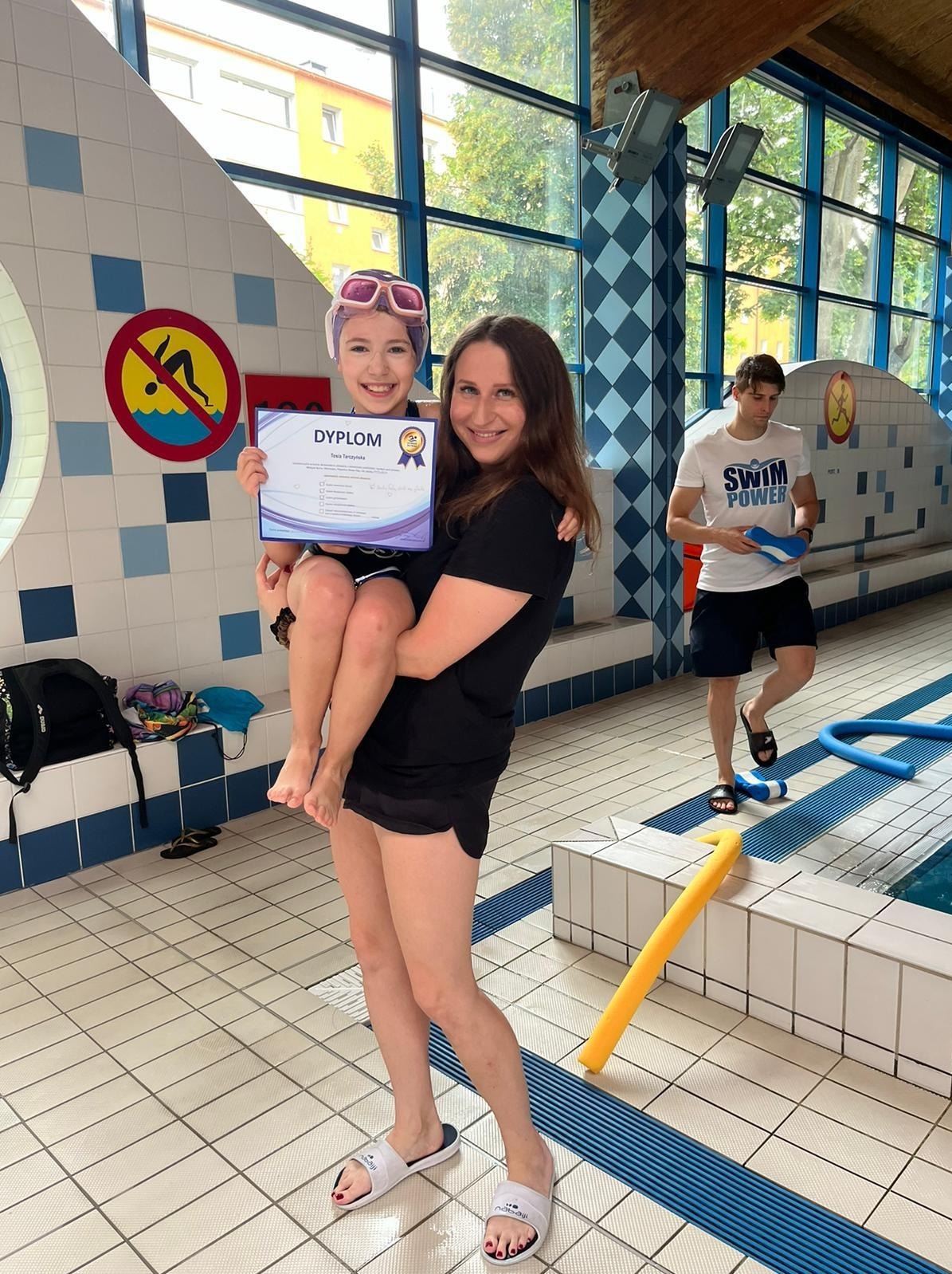 Uśmiechnięta dziewczynka w czepku i okularach pływackich trzyma dyplom ukończenia kursu pływania, niesiona na rękach przez instruktorkę na tle basenu.